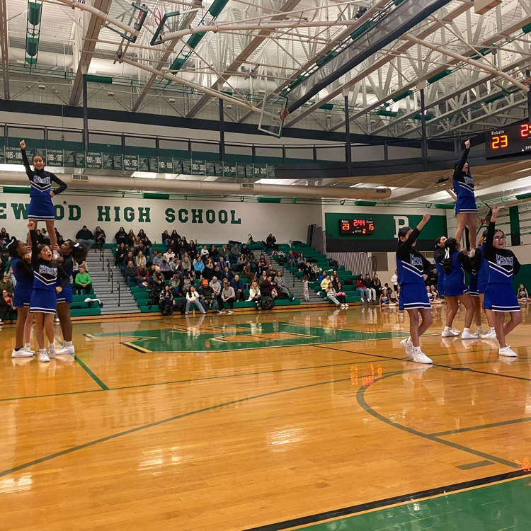 A photo of the Mannheim cheerleading squad performing stunts on the basketball court at Ridgewood High School. The cheerleaders, wearing blue and black uniforms, are executing two lift formations with flyers raised in the air while teammates support them. The background features a crowd of spectators in the bleachers watching the performance during the game.