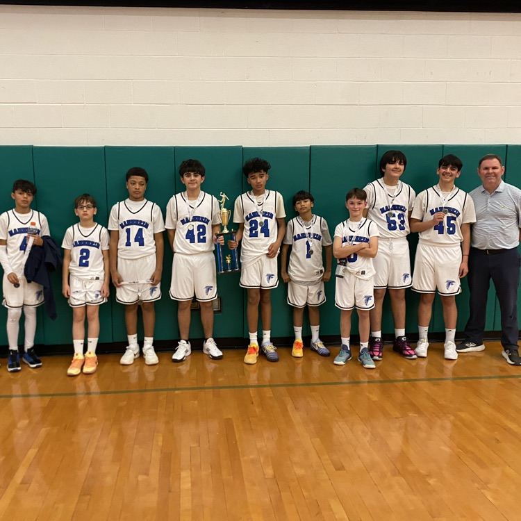 A group photograph of the Mannheim boys' basketball team and their coach posing together on a wooden gymnasium floor. Ten players, wearing white uniforms with blue "MANNHEIM" text, stand in front of a green padded wall, with one player in the center holding a trophy. Their coach stands on the far right, smiling.