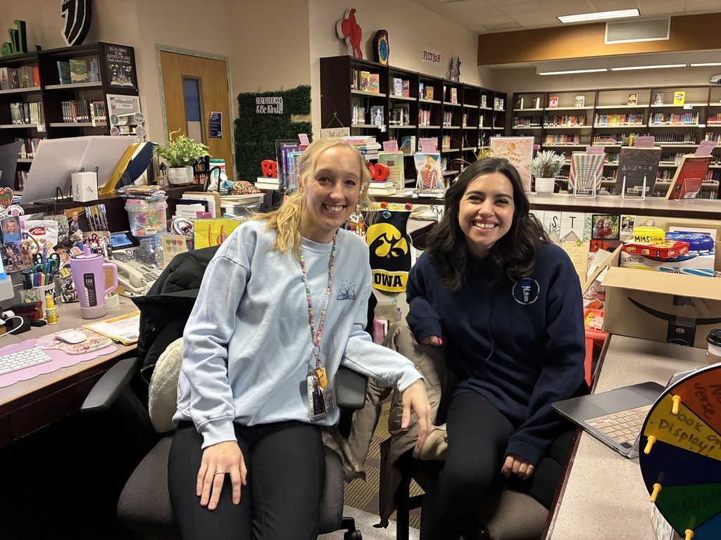 A photo of two smiling staff members seated together at a desk in the school library. The background features bookshelves labeled "Fiction" and various decorations, while the desk is filled with colorful office supplies and an Iowa pennant. The image captures a friendly and welcoming environment in the media center.