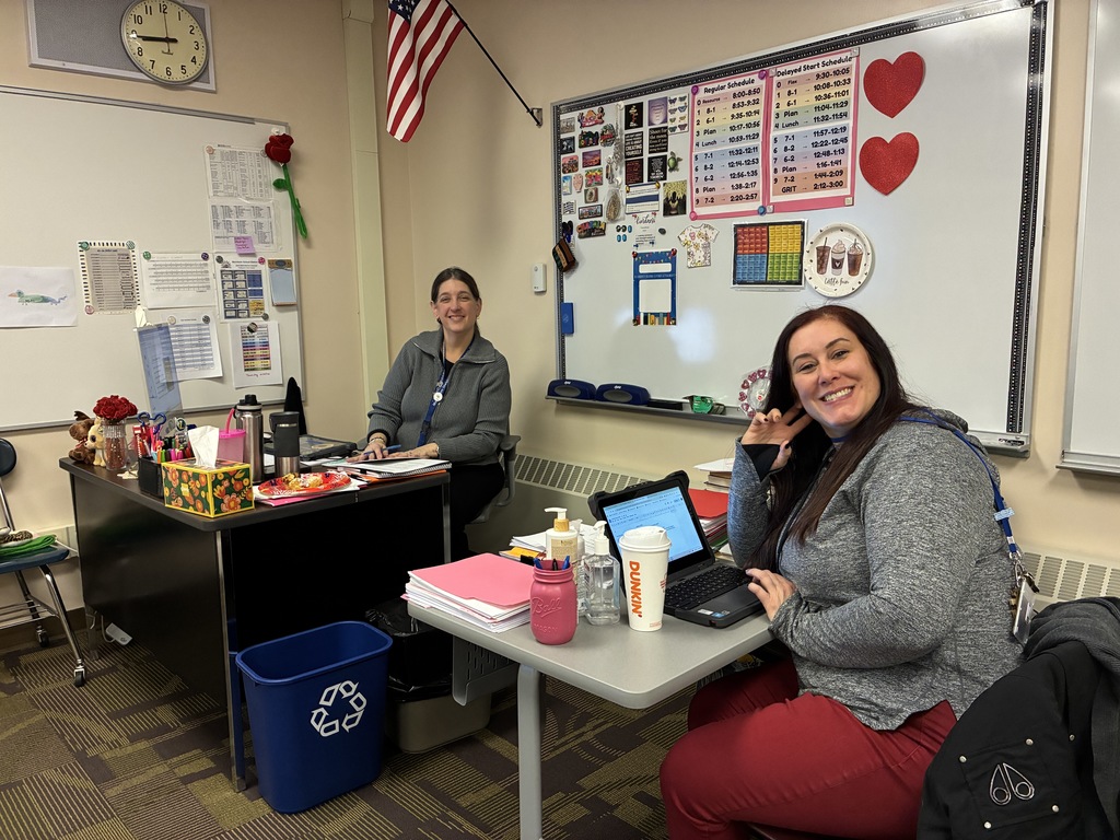 Alt Text A photo of two smiling staff members seated at their desks in a classroom office. The woman on the left sits behind a desk with a computer, while her colleague sits at a separate table with a laptop and a coffee cup, posing with a peace sign. The background features a whiteboard with daily schedules and red heart decorations, creating a cheerful atmosphere.