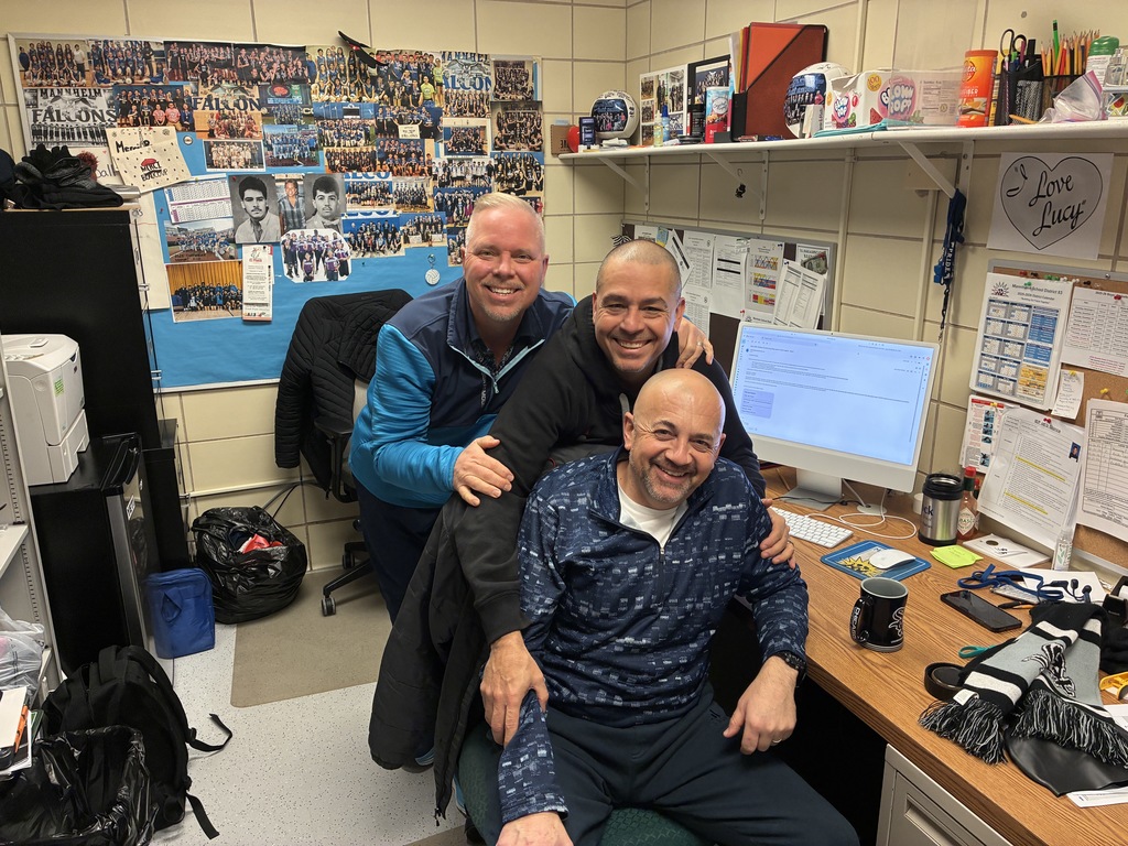 A photo of three smiling staff members posing together in an office. One man is seated at a desk while two colleagues stand behind him, leaning in for a friendly group picture. The office walls are decorated with numerous Mannheim Falcons team photos, calendars, and personal memorabilia, capturing a warm moment of camaraderie and school spirit.