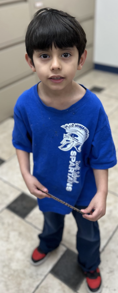 A young student with short dark hair stands in a school office, wearing a blue t-shirt with a white "Scott School Spartans" logo. He is holding a thin, patterned band with both hands, dressed in dark pants and red and black sneakers, with beige file cabinets visible behind him.