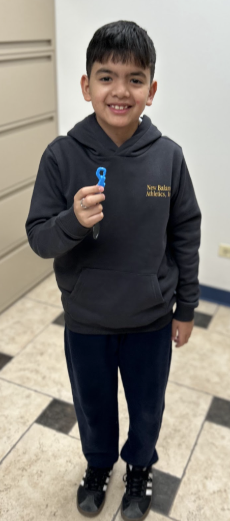 A smiling student wearing a dark "New Balance" hoodie stands in a school office holding a small blue object in his hand.