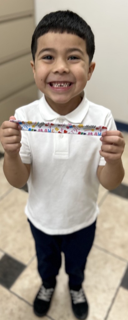 A smiling young student with short dark hair holds up a colorful slap bracelet featuring text like "OMG" and "LOVE" with both hands. He is wearing a white polo shirt, dark pants, and black sneakers, standing on a tiled floor in a school office with beige file cabinets behind him.