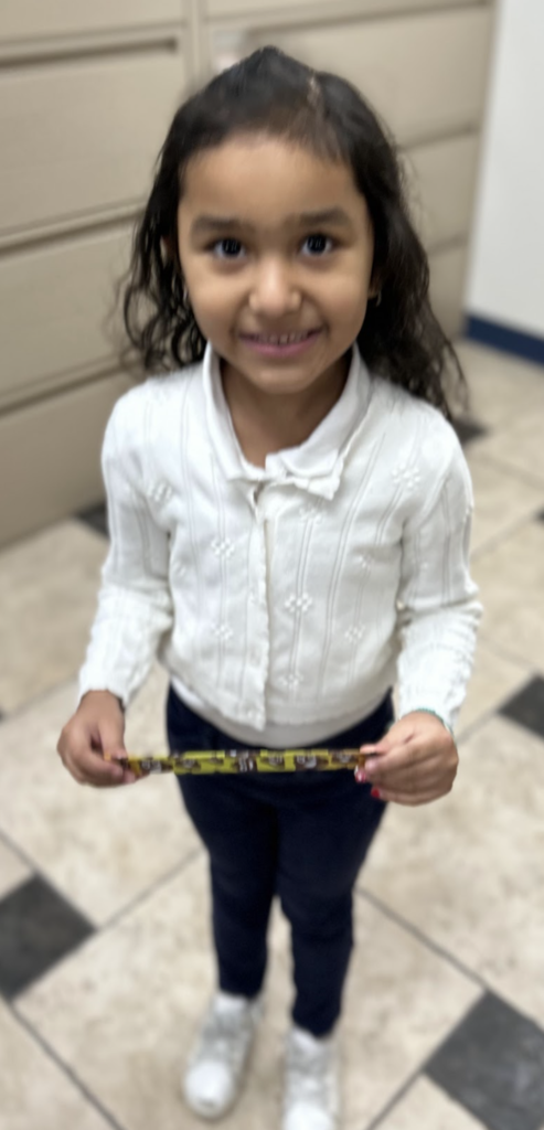A smiling young student with long wavy hair stands in a school office, holding a small yellow and brown object with both hands. She is wearing a white textured cardigan, dark pants, and white sneakers, with beige file cabinets in the background.