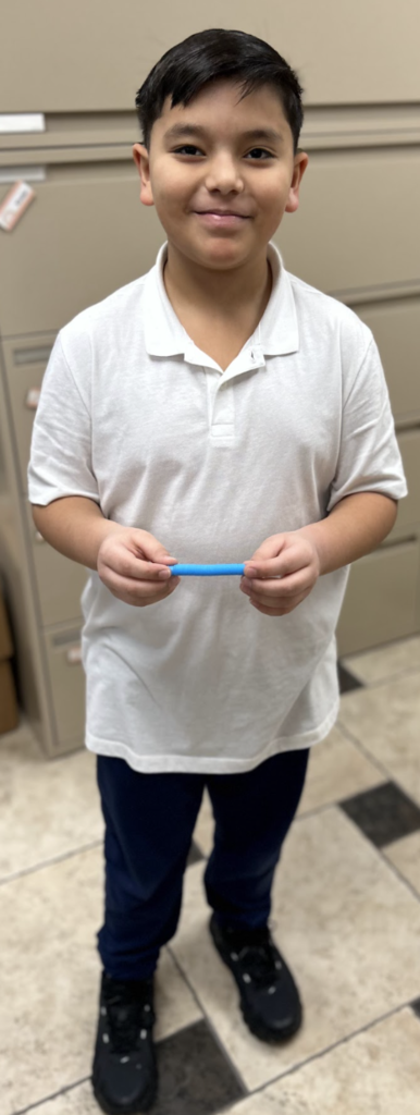 A smiling young student with short dark hair stands in a school office holding a small blue object with both hands. He is wearing a white polo shirt, dark pants, and black sneakers, with beige file cabinets in the background.