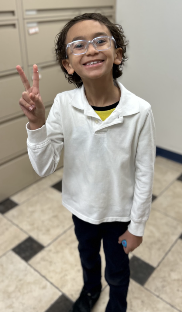 A smiling young student with curly hair and clear glasses stands in the school office, holding up a peace sign with his right hand. He is wearing a white long-sleeved polo shirt and dark pants, with file cabinets visible in the background.