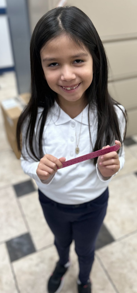 A smiling young student with long dark hair stands in a school office, holding a pink patterned band with both hands. She is wearing a white long-sleeved polo shirt with a necklace and dark pants, with beige file cabinets and a cardboard box visible in the background.