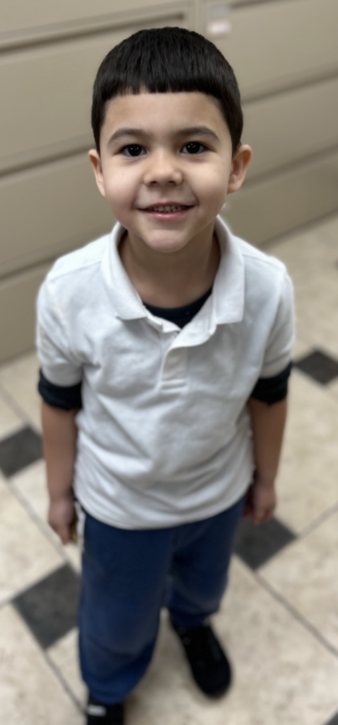 A smiling young student with short dark hair stands in a school office with a tiled floor. He is looking up at the camera wearing a white polo shirt and blue pants, with beige file cabinets behind him.