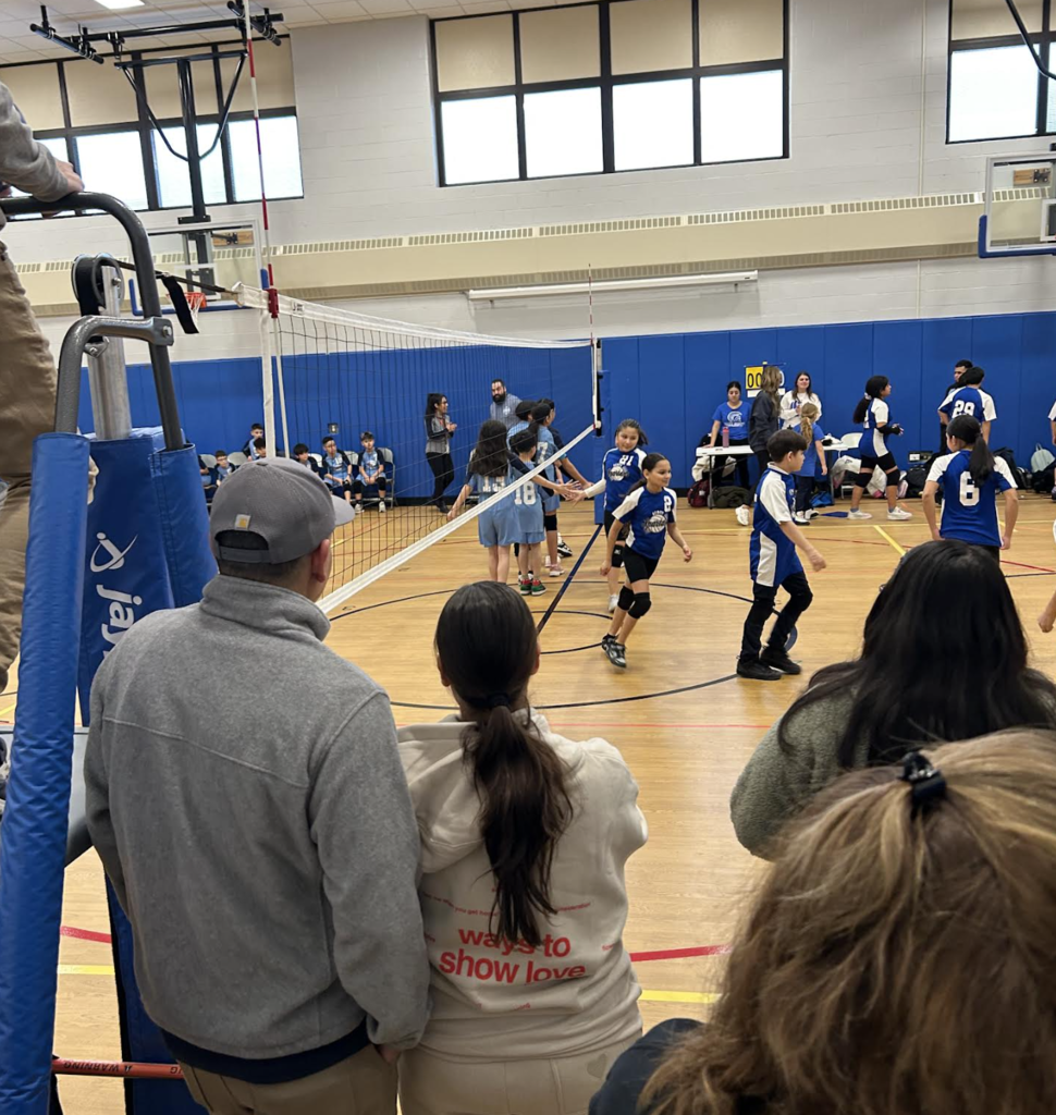 A view from behind spectators watching a youth volleyball game in a gymnasium. Two teams are shaking hands at the net under the supervision of a coach. The foreground shows the backs of several people, including a person in a grey jacket and another in a tan hoodie with red text that reads "ways to show love." A tall blue padded referee stand is visible on the left side of the net.