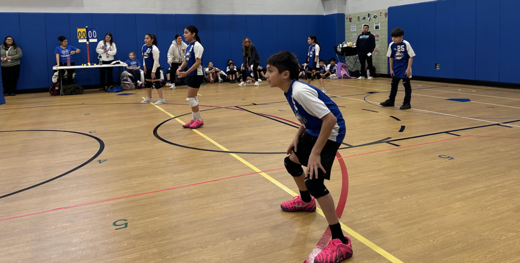 A group of youth volleyball players in blue and white jerseys are positioned on an indoor court during a game. In the foreground, a boy in bright pink sneakers and black knee pads stands in a ready stance, focused on the play. Behind him, other teammates are spread across the wooden court, while spectators and staff sit near a scoreboard along the blue padded gymnasium wall.