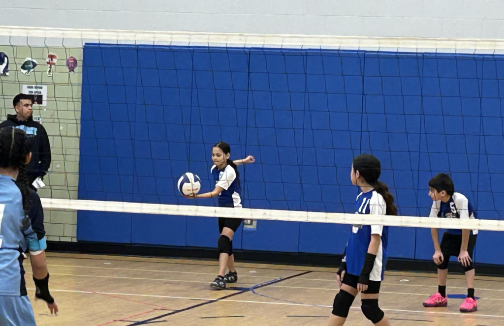 A young volleyball player in a blue and white jersey prepares to serve the ball during an indoor game. She stands mid-court, holding the volleyball in her left hand with her right arm pulled back, ready for an underhand serve. Two teammates in matching uniforms and black knee pads look on from the foreground, while a coach or official stands near the net in the background against a blue padded wall.