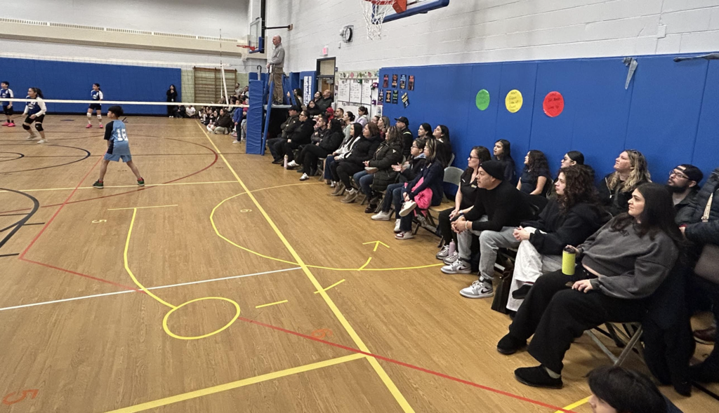 An action-oriented view from the sidelines of a youth volleyball game in a school gymnasium.  A group of spectators sits on folding chairs and benches along a wall with blue protective padding, watching the match intently. On the wooden court, a player in a light blue jersey with the number 18 stands ready near the net, while opposing players in dark blue and white uniforms are visible across the court. A referee stands on an elevated platform near the net, and a basketball hoop is mounted on the wall above the seating area.