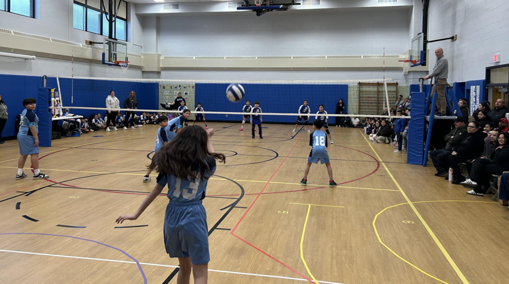 An action-oriented, wide-angle shot of a youth volleyball game in a brightly lit school gymnasium.  In the foreground, a girl wearing a light blue jersey with the number 13 is captured from behind, mid-motion as she serves the ball. The volleyball is frozen in the air just above the net. On her side of the court, three teammates in matching blue uniforms watch the play, while the opposing team in dark blue and white jerseys stands ready across the net.  A referee stands on a raised platform to the right, overseeing the match. Spectators and other players line the perimeter of the court against blue padded walls, and basketball hoops are visible at either end of the gym.