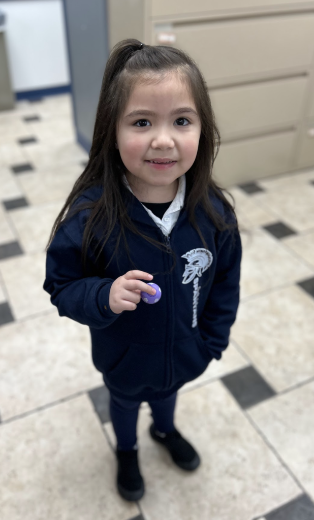 A young girl with long brown hair, partially pulled back, stands in a hallway with checkered floor tiles and smiles at the camera. She is wearing a navy blue zip-up hoodie featuring a "Spartans" logo with a Greek-style helmet on the chest. In her right hand, she holds a small, round purple object that resembles a miniature soccer ball.
