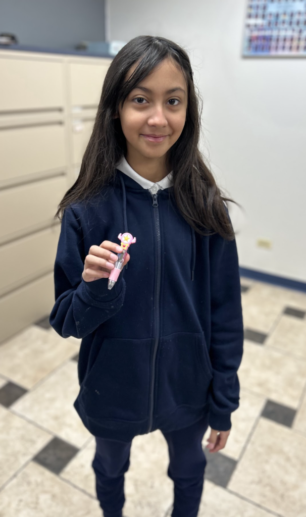 A young girl with long dark hair stands in an indoor setting with tiled floors and filing cabinets, smiling at the camera. She is wearing a dark blue zip-up hoodie over a white collared shirt and is holding a small, pink decorative pen featuring a star-shaped topper.