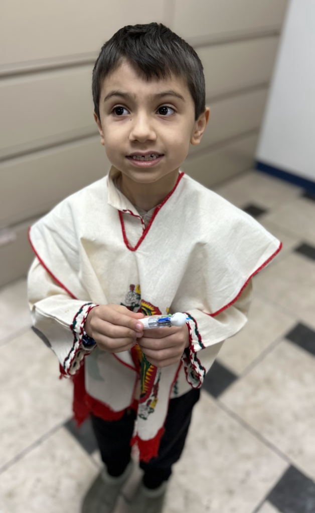A high-angle, full-length shot of a young boy with short dark hair standing on a checkered tile floor. He is wearing a traditional cream-colored Mexican tilma or poncho with red trim and a colorful graphic on the front, paired with dark pants. He is looking slightly off-camera with a gentle smile and is holding a multi-colored retractable pen in both hands.