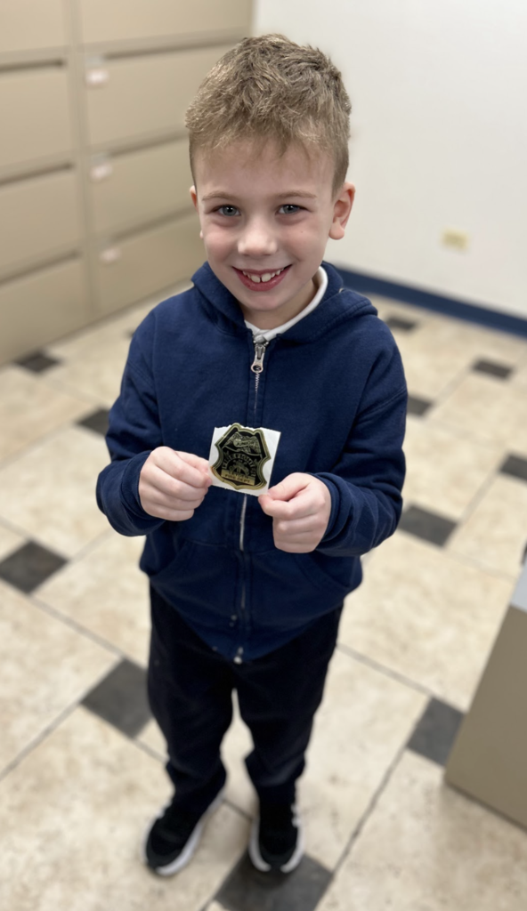 A young boy with short blonde hair and a navy blue zip-up hoodie stands in a tiled hallway, smiling at the camera. He is holding up a gold and black sticker shaped like a fire department badge.