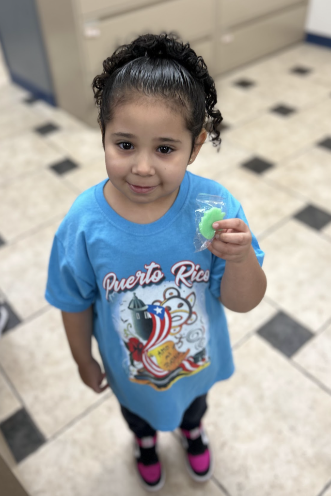 A young girl with dark, curly hair tied back stands on a checkered floor, smiling at the camera. She is wearing a bright blue T-shirt featuring a colorful "Puerto Rico" graphic with a flag and a tropical design. In her right hand, she holds up a small, green, leaf-shaped object encased in a clear plastic wrapper.