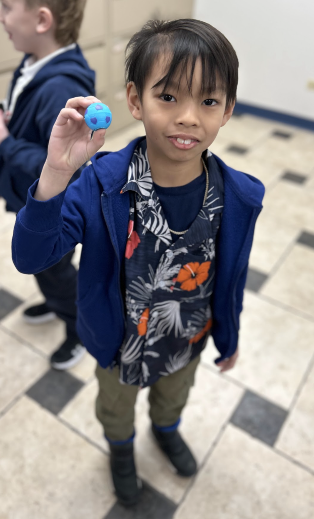 A high-angle, full-length shot of a young boy with dark hair standing on a checkered tile floor. He is wearing a blue zip-up hoodie over a navy blue floral shirt with orange flowers, olive green pants, and black shoes. He is smiling and holding up a small blue and purple ball in his right hand. Another child is partially visible in the background.