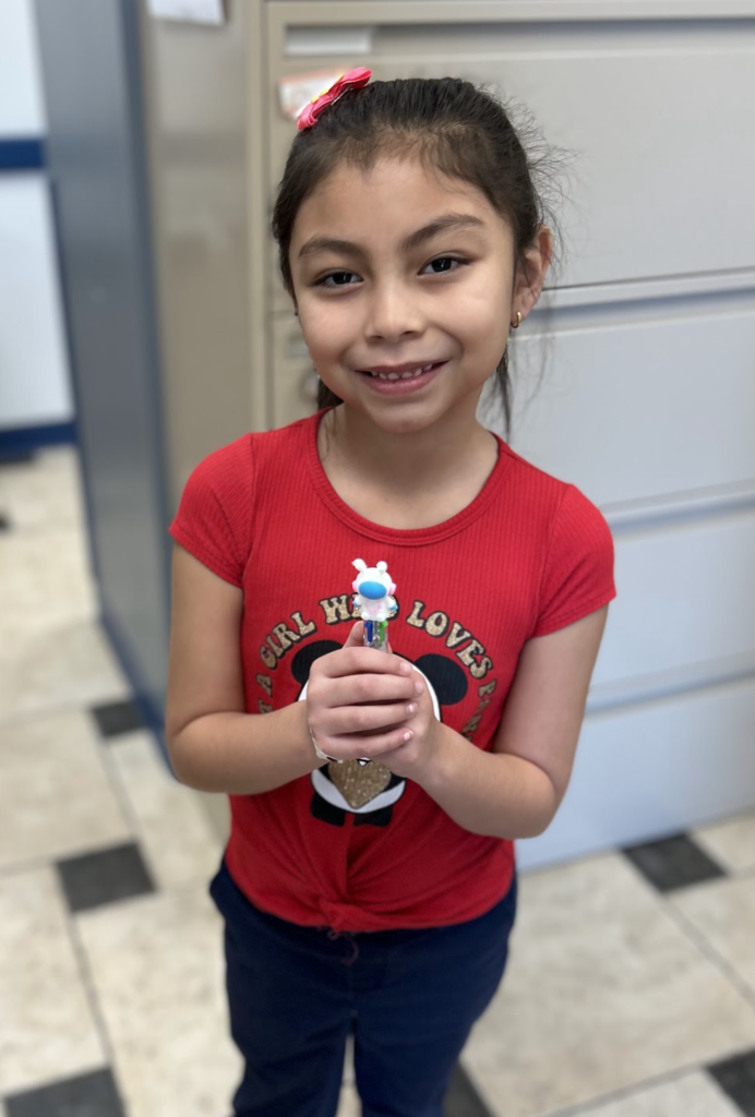 A medium-angle shot shows a smiling young girl with dark hair tied back and a pink bow clip. She is wearing a red short-sleeved shirt with a Mickey Mouse graphic and the words "A girl who loves," paired with dark navy pants. Standing on a checkered tile floor in front of a beige filing cabinet, she holds a white multi-colored retractable pen with a small character topper in both hands.