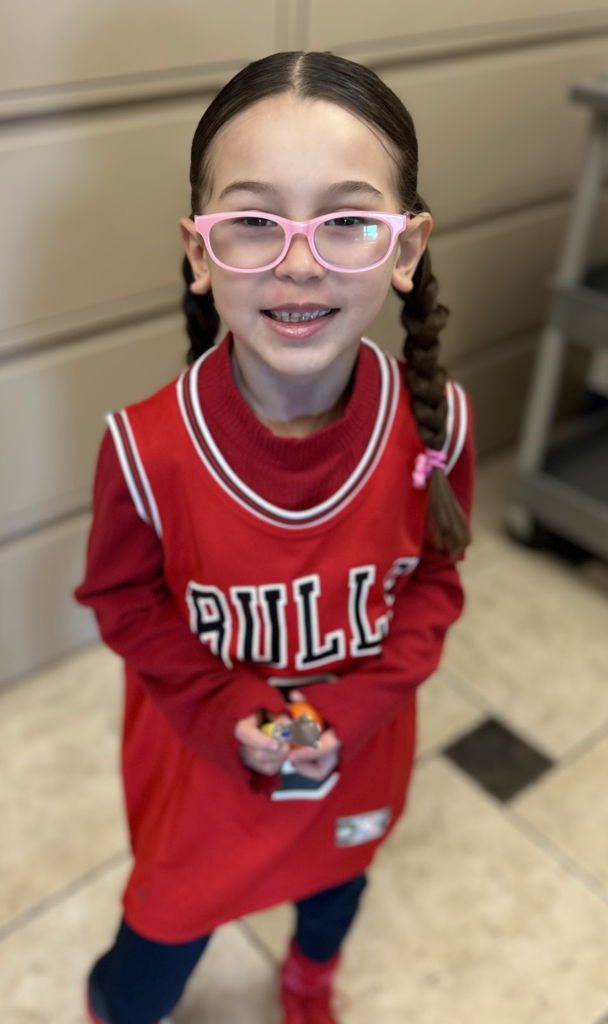 A high-angle, medium shot of a smiling young girl with long dark hair styled in two braids. She is wearing pink-rimmed glasses and a red Chicago Bulls basketball jersey over a matching long-sleeved red shirt, paired with dark leggings. She is standing on a checkered tile floor and holding a small object in both hands.