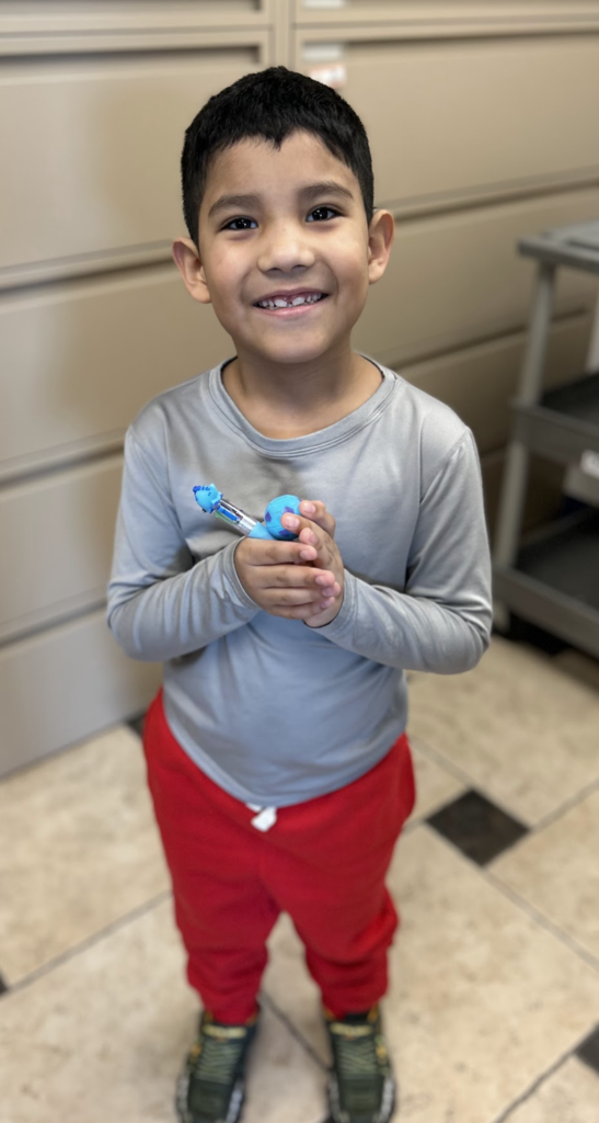 A high-angle, full-length shot of a smiling young boy with short dark hair standing on a checkered tile floor. He is wearing a long-sleeved grey shirt, bright red sweatpants, and dark sneakers with yellow laces. He is holding a blue multi-colored retractable pen and a small blue ball together in both hands.