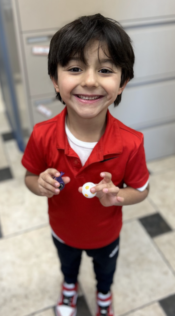 A high-angle, medium shot of a young boy with dark hair smiling widely while standing on a checkered tile floor. He is wearing a red short-sleeved polo shirt over a white t-shirt, dark pants, and red, white, and black sneakers. He is holding a small white ball with yellow spots in one hand and a purple object in the other.