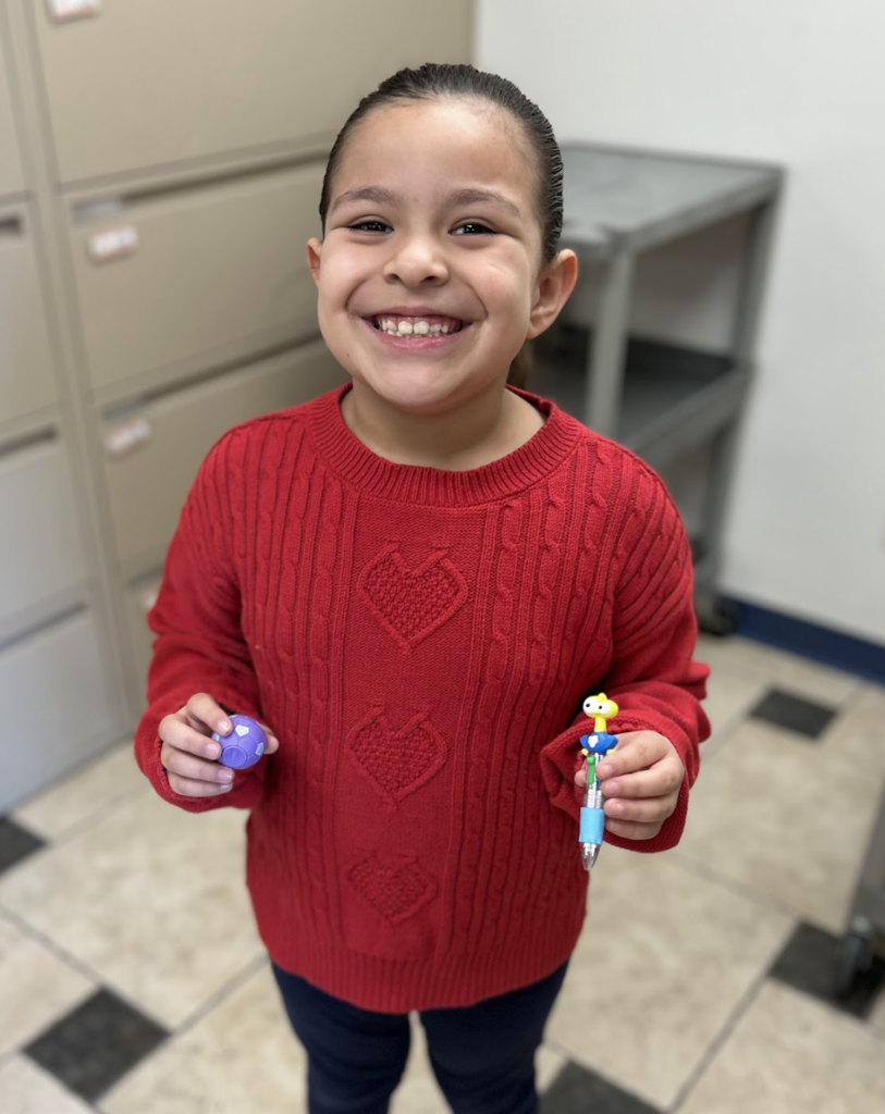 A high-angle, medium shot of a young girl with dark hair pulled back into a ponytail, smiling broadly while standing on a checkered tile floor. She is wearing a vibrant red cable-knit sweater featuring three vertical heart patterns down the center, paired with dark pants. In her right hand, she holds a small purple ball, and in her left, she displays a multi-colored retractable pen topped with a yellow character figure.