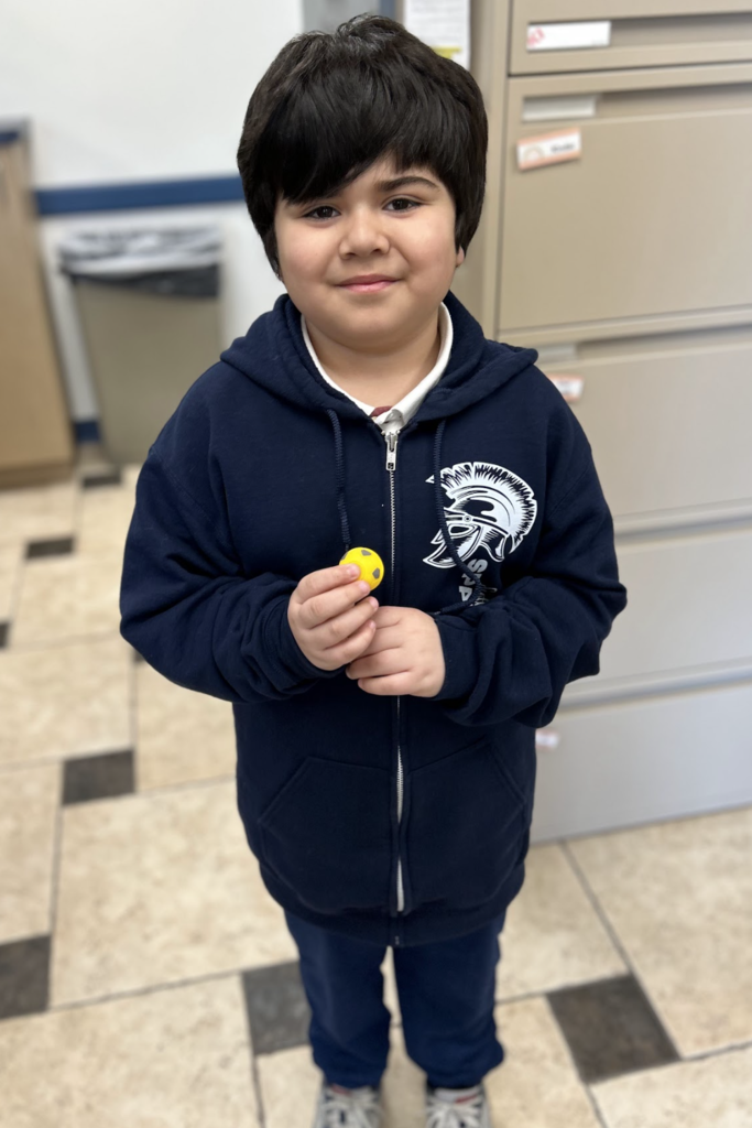A high-angle, medium shot of a young boy with dark, thick hair smiling as he stands on a checkered tile floor. He is wearing a navy blue zip-up hoodie over a white collared shirt and navy pants; the hoodie features a white Spartan logo on the left chest. He is holding a small yellow ball in both hands.