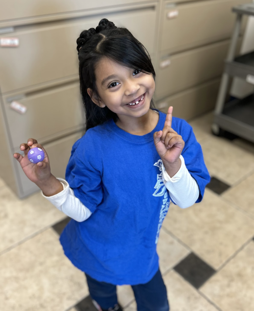 A high-angle shot of a young girl with long dark hair, styled with a braided crown, smiling broadly and missing a few front teeth. She is wearing a bright blue short-sleeved t-shirt over a white long-sleeved shirt and dark pants. Standing on a checkered tile floor, she holds a small purple and white ball in her right hand while pointing upward with her left index finger.