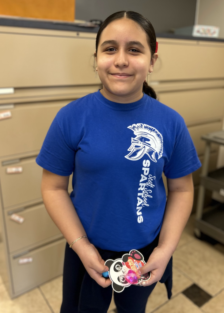 A medium-angle, waist-up shot of a smiling girl with dark hair tied back. She is wearing a bright blue t-shirt with a white Scott School Spartans logo on the left side, paired with dark pants. Standing on a checkered tile floor in front of beige filing cabinets, she holds a panda-themed card and a small pink prize in both hands.