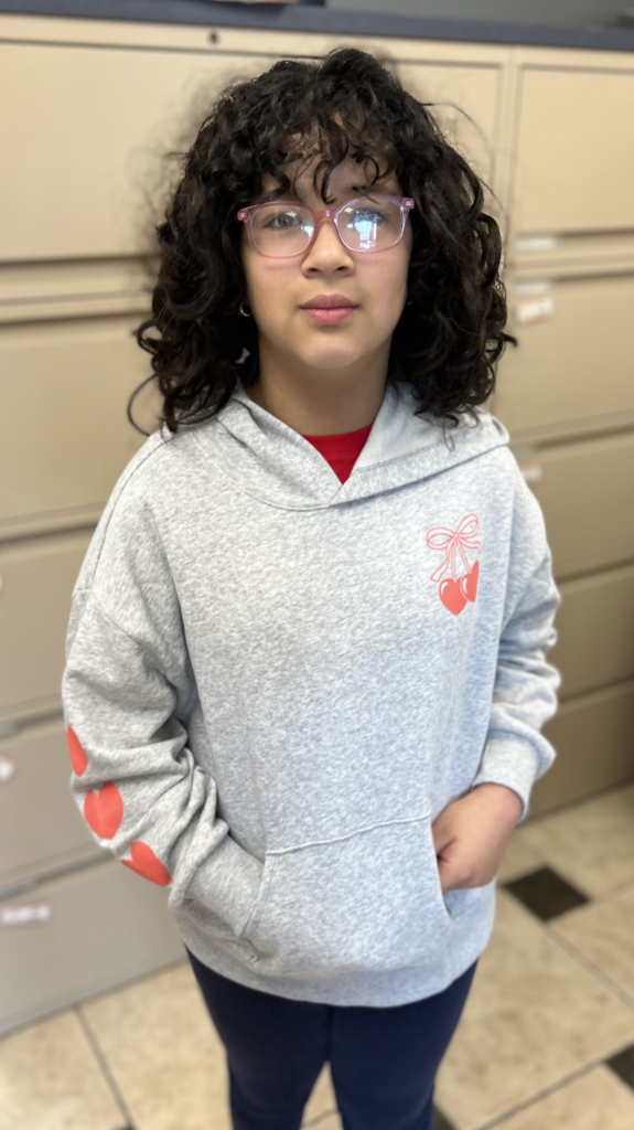 A medium-angle, full-length shot of a girl with dark, curly hair and pink-framed glasses standing on a checkered tile floor. She is wearing a light grey hooded sweatshirt featuring a small graphic of two red hearts tied with a bow on the chest and a series of red hearts down the right sleeve, paired with dark blue pants. She is standing with her hands in her sweatshirt pocket.