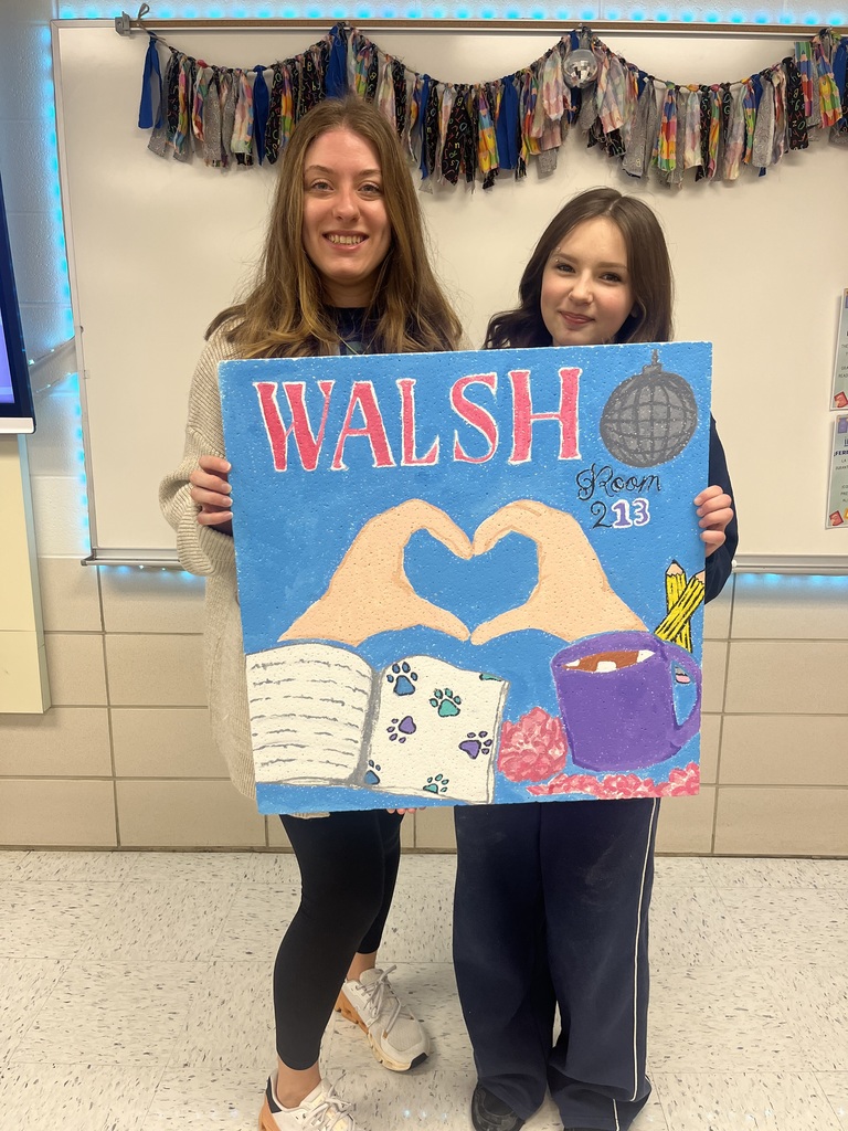 Alt Text: Two women stand in a classroom holding a large, hand-painted ceiling tile. The tile has a bright blue background with the name “WALSH” in large pink letters and “Room 213” written on the right side next to a disco ball. In the center, two hands form a heart shape above an open book decorated with colorful paw prints. A purple mug filled with a warm drink and yellow pencils sit on the right side of the design, with pink decorative accents along the bottom. A garland hangs across the whiteboard behind them.