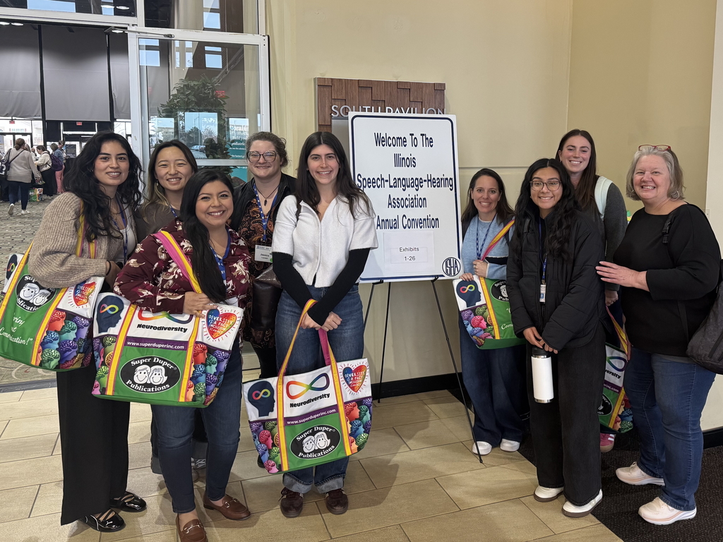 A group of nine smiling women pose together at the Illinois Speech-Language-Hearing Association (ISHA) Annual Convention. They are gathered around a white welcome sign on an easel in a convention center hallway. Several women are holding large, colorful tote bags from Super Duper Publications that feature the neurodiversity infinity symbol and vibrant brain graphics. The group is dressed in casual professional attire, with many wearing convention name badges on blue lanyards.