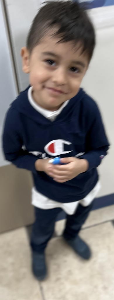 A young boy with short, dark hair stands smiling indoors. He is wearing a navy blue Champion brand hoodie over a white shirt, paired with dark pants and dark shoes. He is holding a small blue object in his hands. The photo has a slight motion blur and is taken from a high angle.