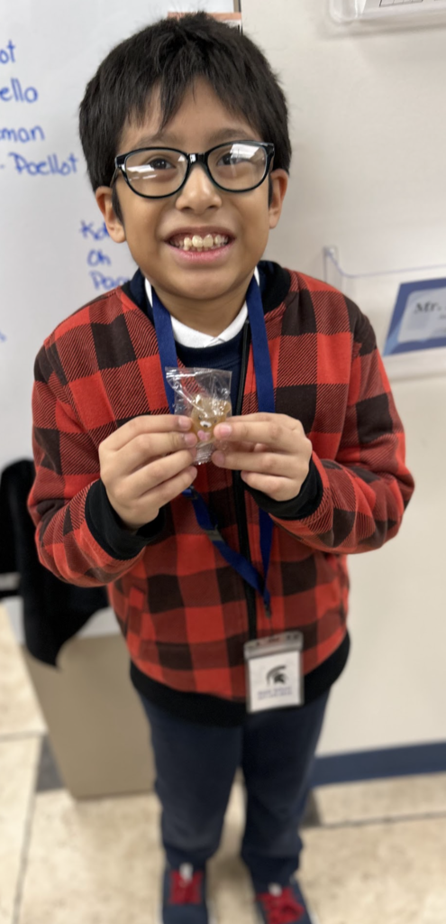A smiling young boy with glasses wears a red and black buffalo plaid zip-up jacket and a blue lanyard with an identification badge. He stands in a classroom setting, holding a small, clear-wrapped bear-shaped treat in both hands. In the background, a white whiteboard features handwritten names, and a tiled floor is visible below.