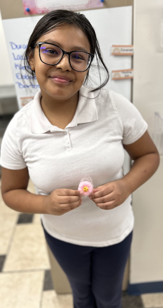 A smiling girl with dark hair and black-rimmed glasses poses in a classroom, wearing a white short-sleeved polo shirt and dark pants. She is holding a small, pink flower-shaped item with a yellow smiley face in the center, wrapped in clear plastic. In the background, a whiteboard with handwritten blue text and several name tags are visible.