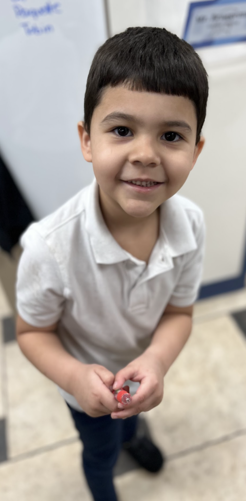 A young boy with short, dark hair and a wide smile stands indoors, looking directly at the camera. He is wearing a white short-sleeved polo shirt and dark pants, and he is holding a small red and clear object in his hands. The photo is taken from a slightly high angle against a blurred background that includes a whiteboard and tiled flooring.