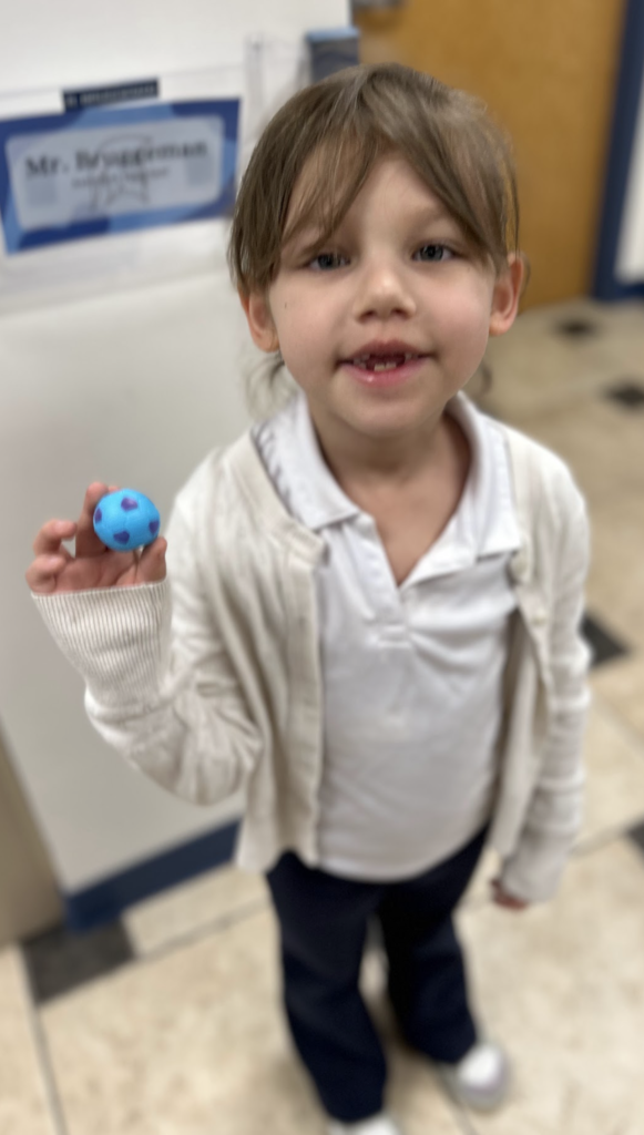 A young girl with brown hair smiling in a school office, wearing a white polo shirt and cream cardigan. She holds up a small, light-blue ball with purple spots. In the background, a nameplate for "Mr. Bruggeman" is visible on the wall.