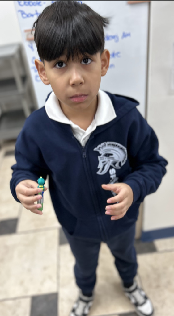 A young boy with dark hair and brown eyes stands in a classroom, looking directly at the camera with a neutral expression. He is wearing a navy blue zip-up hoodie over a white collared shirt, and dark pants. In his right hand, he holds a small, green and blue toy or pen. The background shows a white door or partition with blue handwriting and a tiled floor.