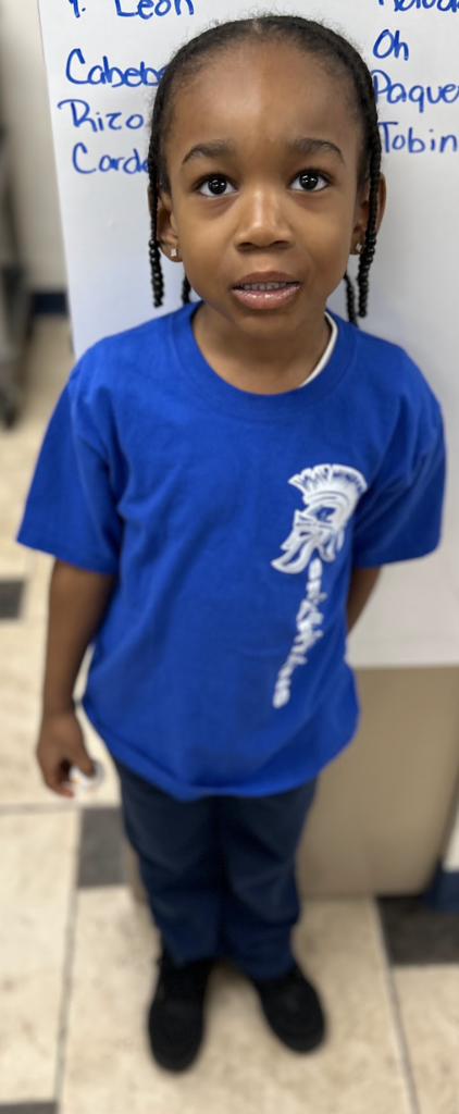 A young child with dark hair in braided pigtails stands in a classroom, looking slightly upward toward the camera. They are wearing a bright blue t-shirt with a white graphic of a Spartan helmet on the left side, dark pants, and black shoes. Behind them is a white board with names written in blue marker, and the floor is a light-colored tile with dark accents.