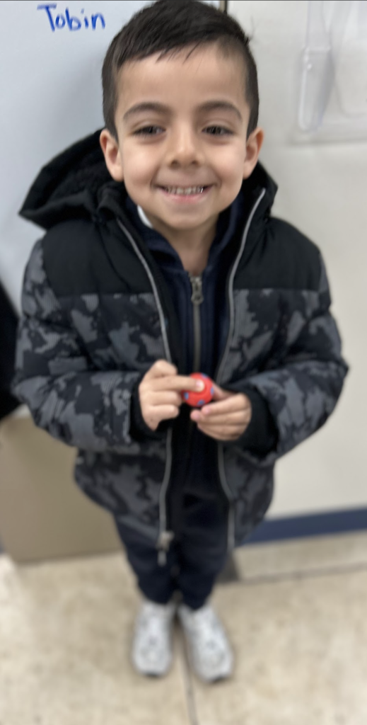 A young boy with short dark hair smiling in a school office, wearing a grey and black patterned winter jacket over a navy zip-up hoodie. He is holding a small red ball with blue spots. In the background, the name "Tobin" is written on a white surface.