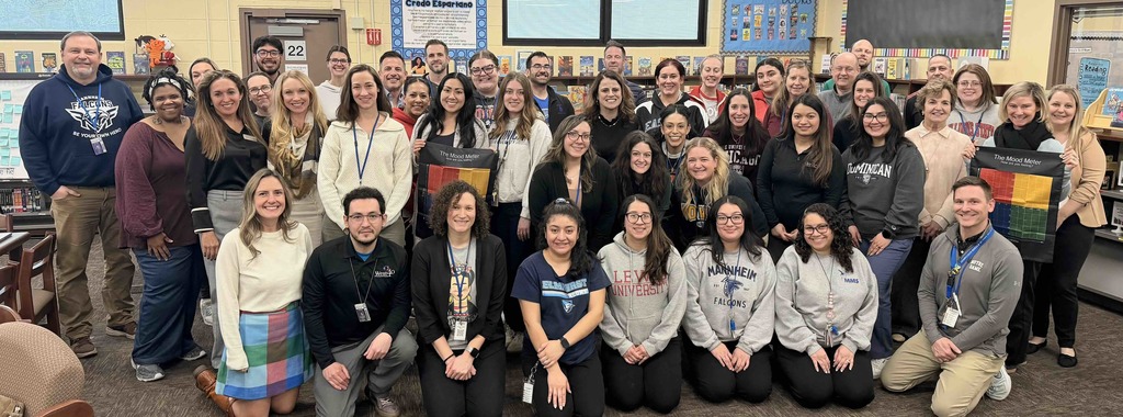 A large group of Mannheim School District staff members pose for a group photo in a school library after a professional development session. The diverse team of educators and administrators are smiling, with some holding "The Mood Meter" posters. Many staff members are wearing school-affiliated apparel, including Mannheim Falcons and various university sweatshirts, showcasing a collaborative and positive school culture.