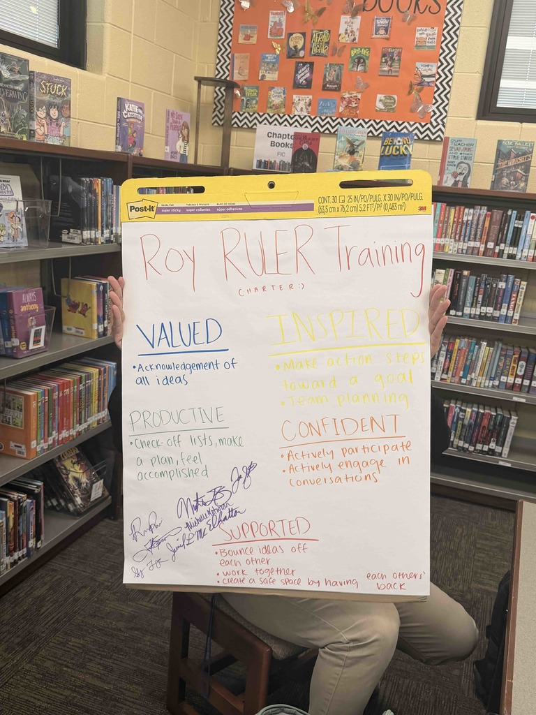 A large Post-it easel pad titled "Roy RULER Training Charter" is held up in a school library, surrounded by bookshelves. The poster displays colorful handwritten notes on group values, including "Valued," "Productive," "Inspired," "Confident," and "Supported," with corresponding action items like "Acknowledgement of all ideas" and "Work together." Several staff signatures are visible in the bottom left corner, signifying team agreement.