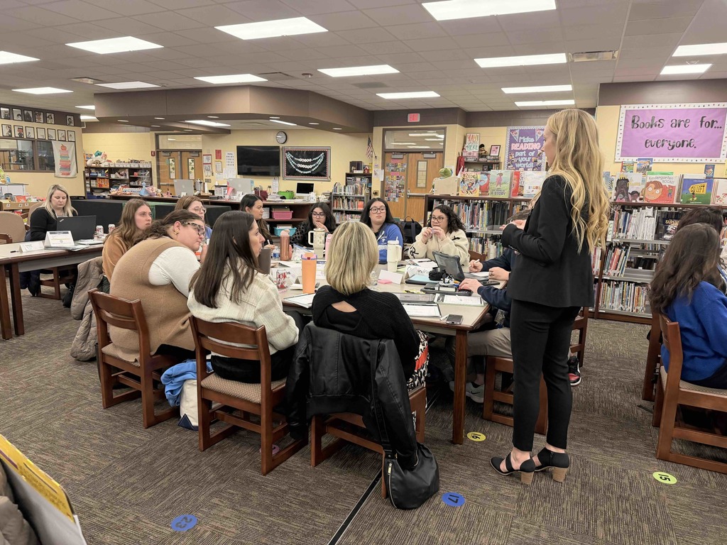 A group of educators and staff participate in a professional development workshop in a school library. A facilitator in a black suit stands at the head of a large table, addressing several seated participants who are engaged in discussion and taking notes. Bookshelves and a sign reading "Books are for everyone" are visible in the background.