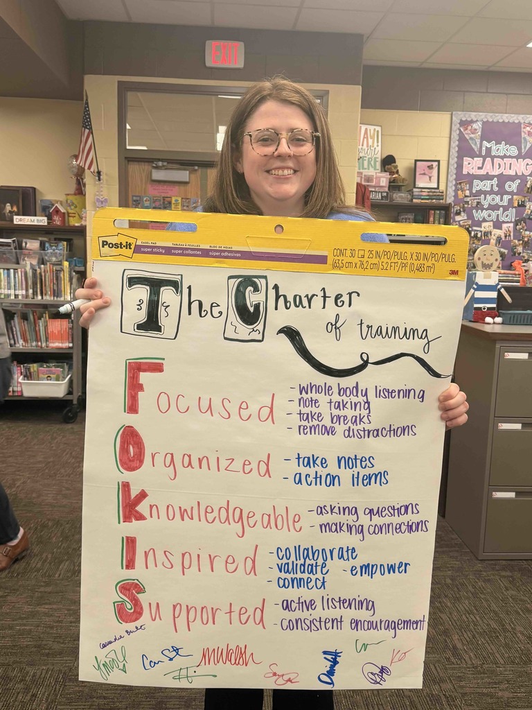 A smiling staff member holds up a hand-drawn poster titled "The Charter of Training" in a school library. The poster uses the acronym "FOKIS" to highlight team values: Focused, Organized, Knowledgeable, Inspired, and Supported. Each value includes specific goals like "whole body listening," "action items," and "consistent encouragement," with several colorful staff signatures at the bottom of the page.