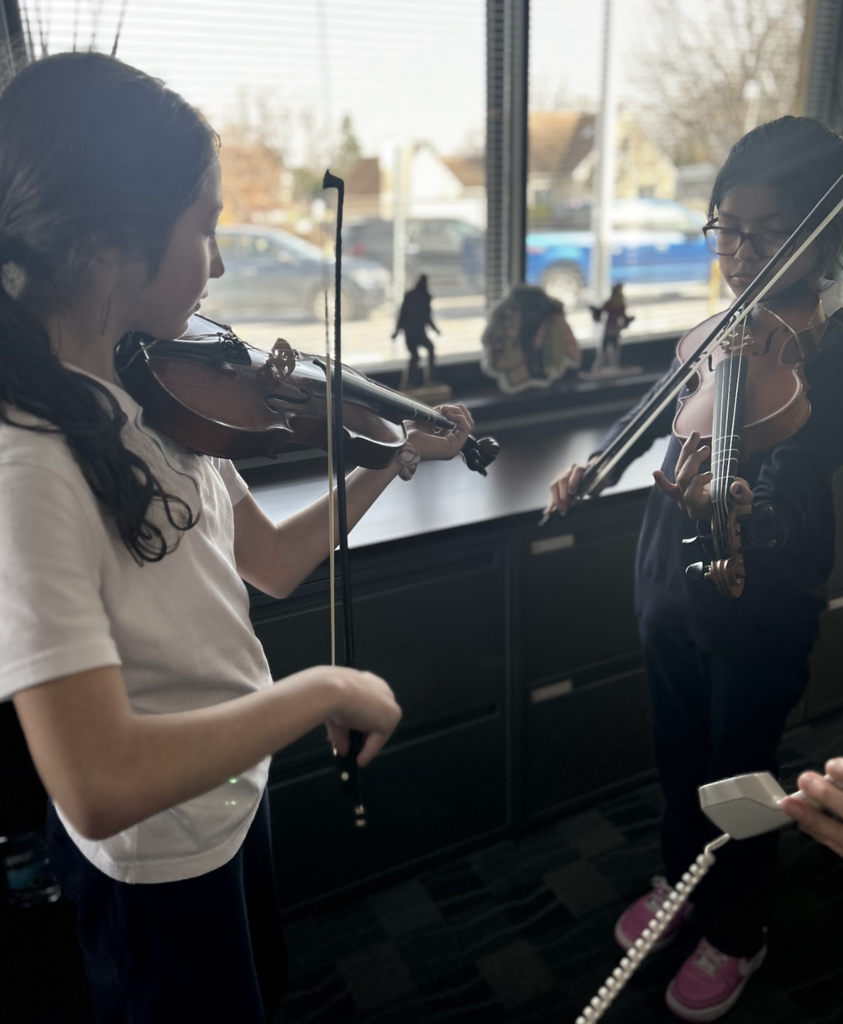 Two young girls stand indoors near a large window, focused on playing their violins. The girl on the left, wearing a white t-shirt, is shown in profile, while the girl on the right wears glasses and a dark sweater. Natural light streams in from the window behind them, which overlooks a street with parked cars. The scene captures a quiet moment of musical practice and concentration.