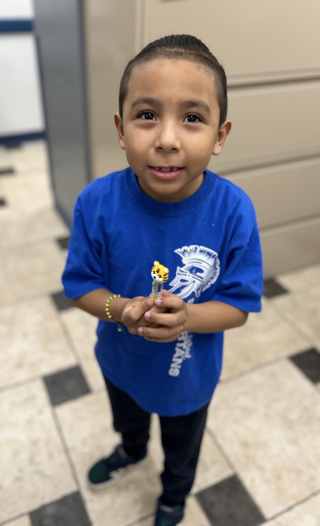 A medium shot shows a young boy with dark hair and a buzz cut, looking upward toward the viewer with a smile. He wears a bright blue t-shirt with a white graphic of a Spartan helmet and "Spartans" text, dark blue pants, and green and black sneakers. He is holding a small, yellow tiger toy with both hands at his chest. The boy stands in a school hallway with a beige tiled floor and black square accents, a brown door to his right, and blue cabinets to his left.
