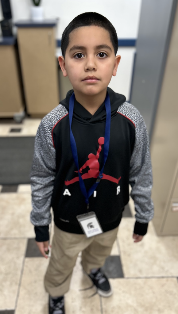 A young boy with a serious expression, wearing a black Jordan hoodie with patterned grey sleeves and a red logo, dark blue lanyard, tan pants, and sneakers, stands in a room with a tiled floor and a blurred background.